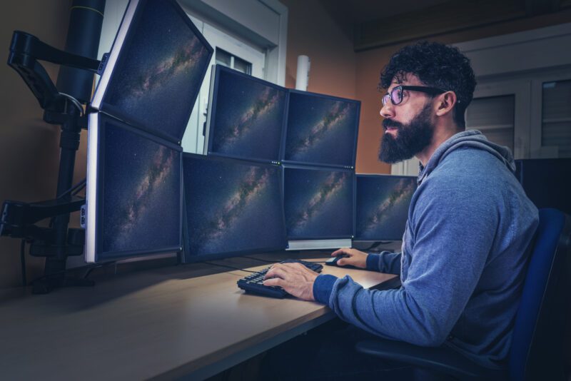 Astronomer in control panel room multi screen sitting profile with beard- Careers for Students Who Love Math