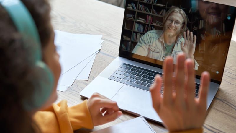 Hispanic teen girl school college student distance learning waving hand studying with online teacher on laptop screen. Elearning zoom video call, videoconference class with tutor. Over shoulder view. (Hispanic teen girl school college student distance