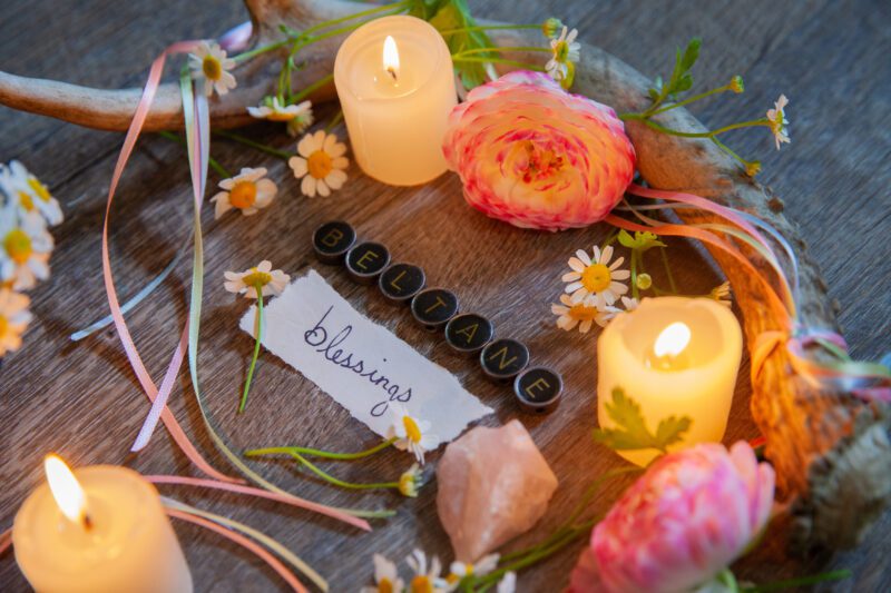 Altar set for the Mid-Spring Holiday: Beltane. The altar features deer antler, candles, pastel colored ribbons, chamomile flowers, Ranunculus blooms, and vintage type writer keys and handwritten script to read "Beltane Blessings."