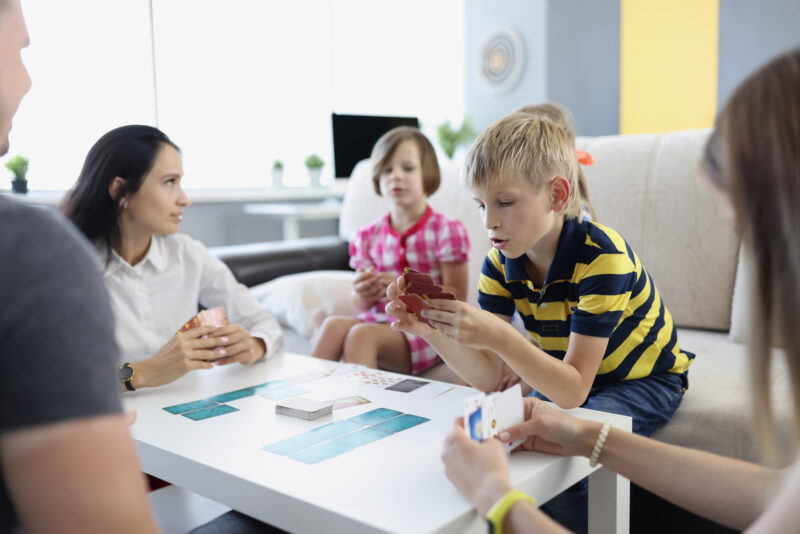 A group of children and adults sit around a table playing cards.