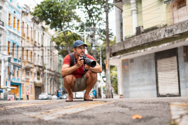 A man is seen crouching down in a street while holding a professional camera. 