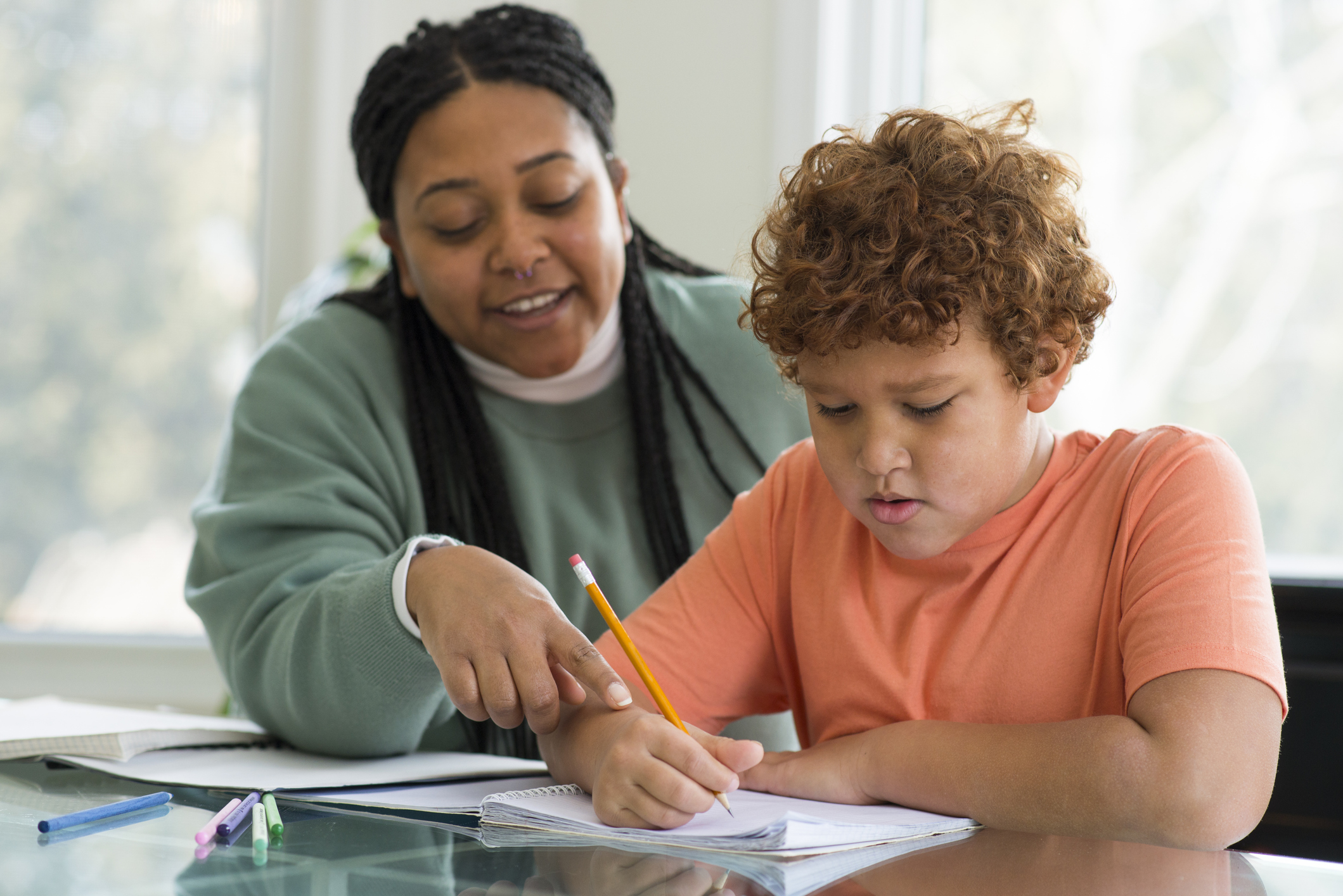 Paraprofessional is smiling gently as she is helping her elementary age student with his school work at a desk. She is pointing at a mistake on the page and he is using a pencil to correct the work in the notebook.