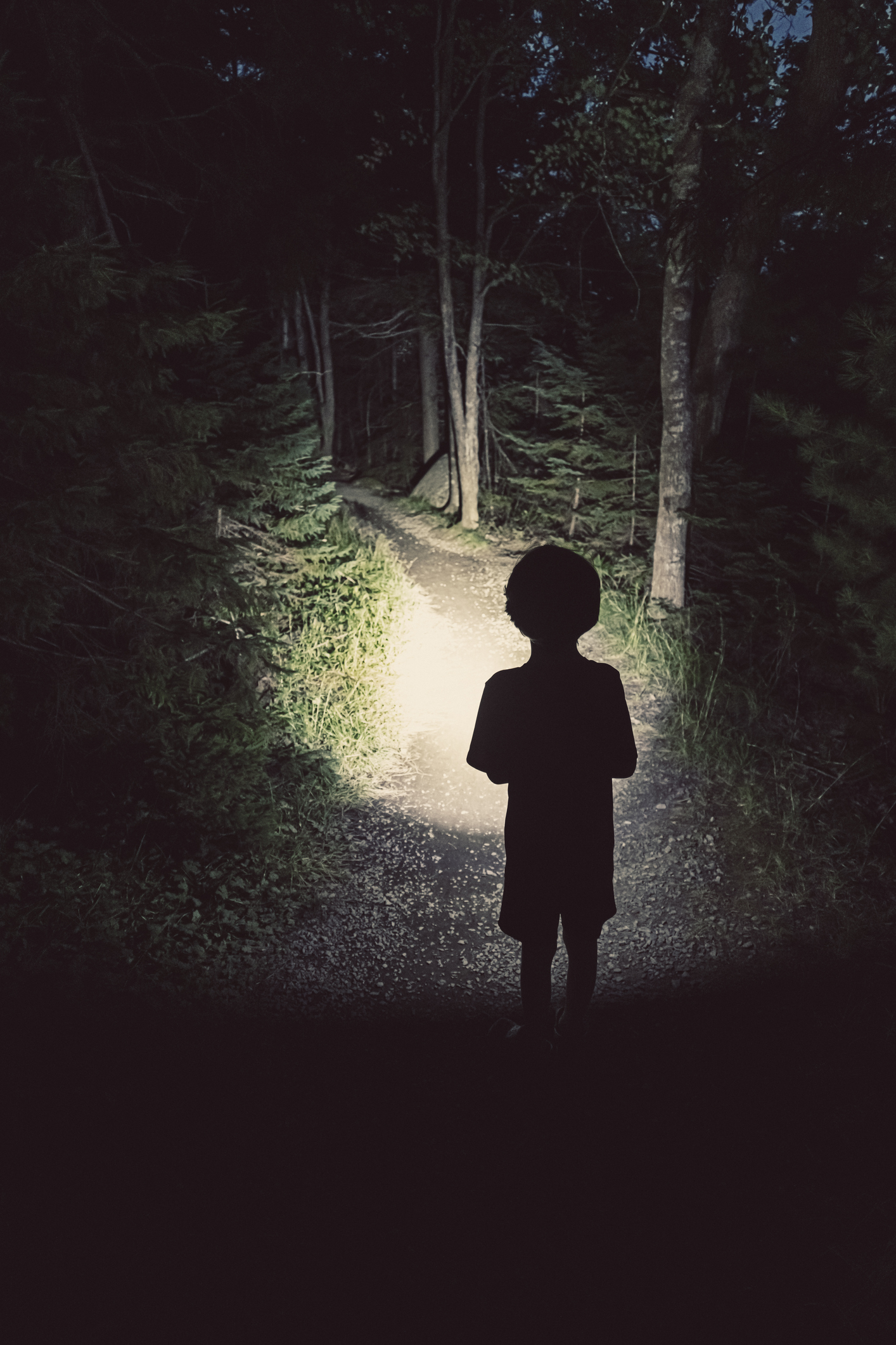summer camp activities like flashlight tag can be played at night. A little boy stands in the dark holding a flashlight into the woods.
