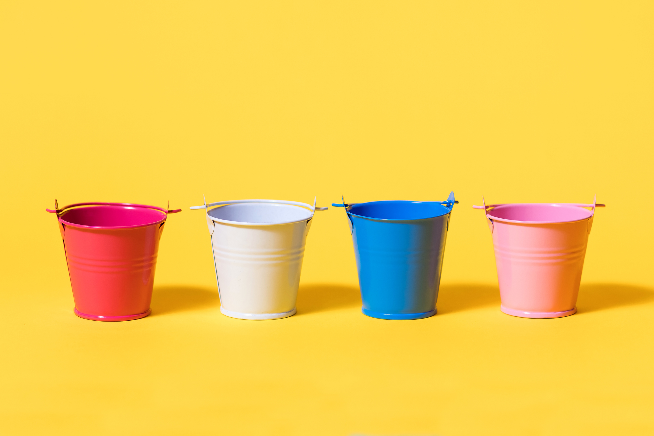 Four multi-colored toy buckets on a yellow background. White blue pink and red bucket standing in a row. Concept of future renovation or start of harvesting or planting crops