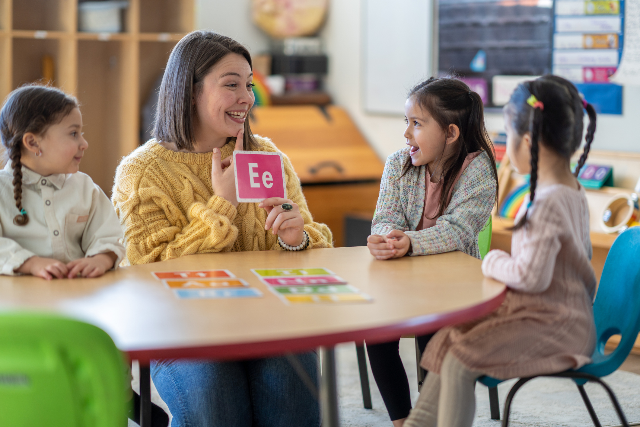 A Kindergarten teacher sits with a small group of students as she teaches them the different sounds each letter of the alphabet makes. Each of the students are dressed casually and paying close attention to their teacher.