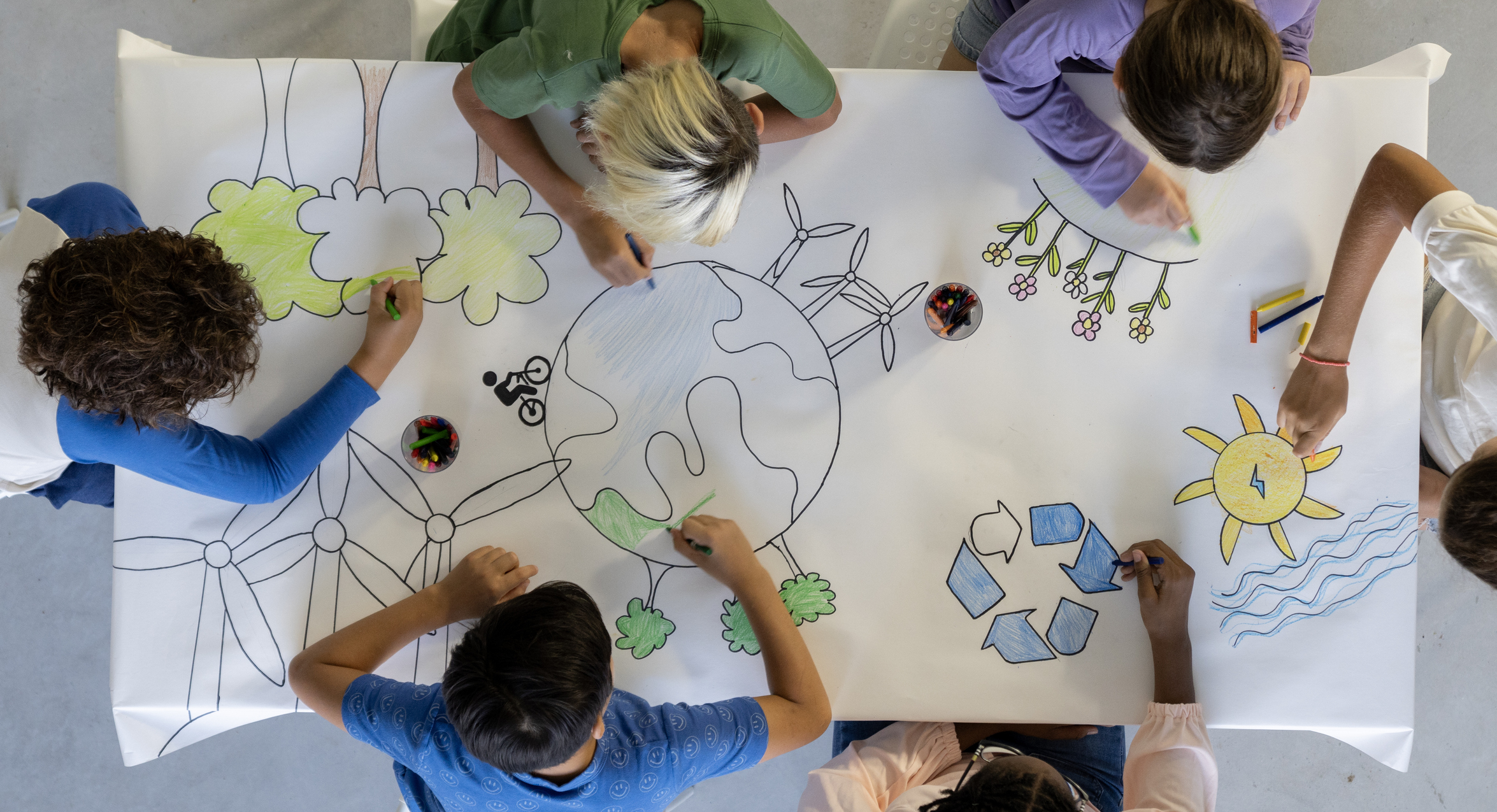 A small group of school aged children sit around a desk as they work together to colour in a drawing. They are each dressed casually and are focused on the task.