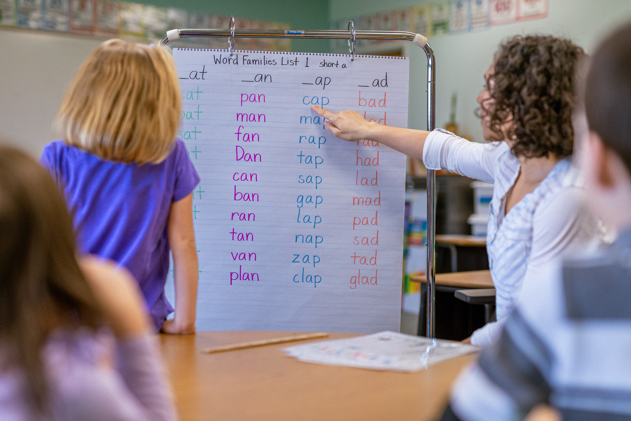 A young teacher of Eurasian ethnicity points to a lesson board as a student stands, learning in an elementary school classroom.