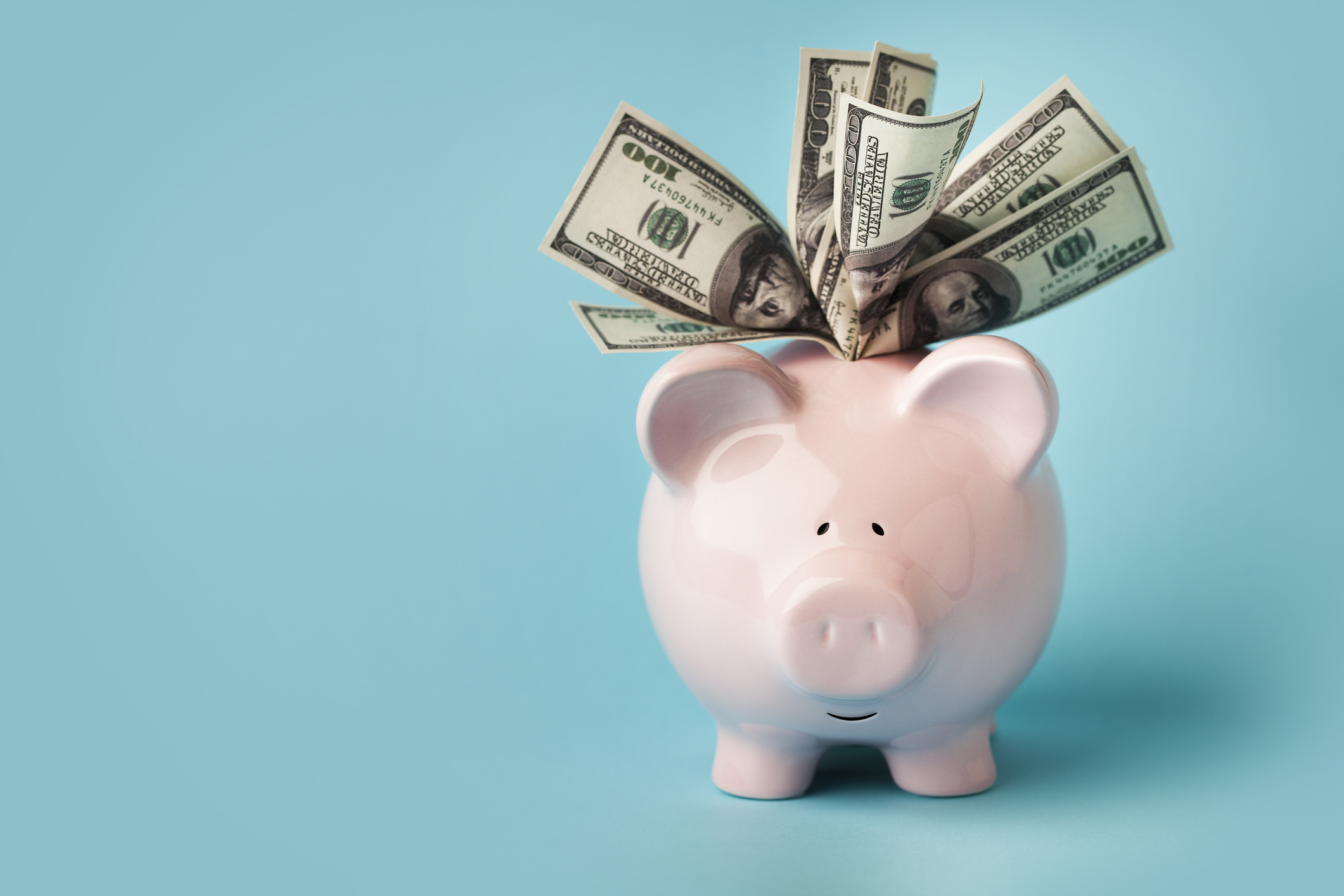 A smiling pink piggybank stuffed with $100 dollar bills, on blue background.