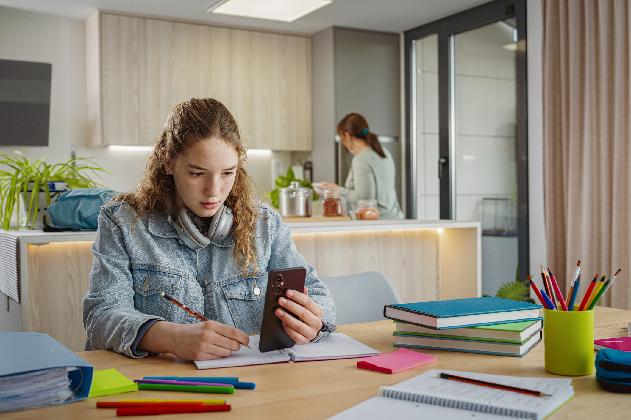 Schoolgirl using smartphone to watch online tutoring lesson. She is sitting at dining room while her mother is cooking at background. High resolution 42Mp studio digital capture taken with SONY A7rII and Zeiss Batis 40mm F2.0 CF lens