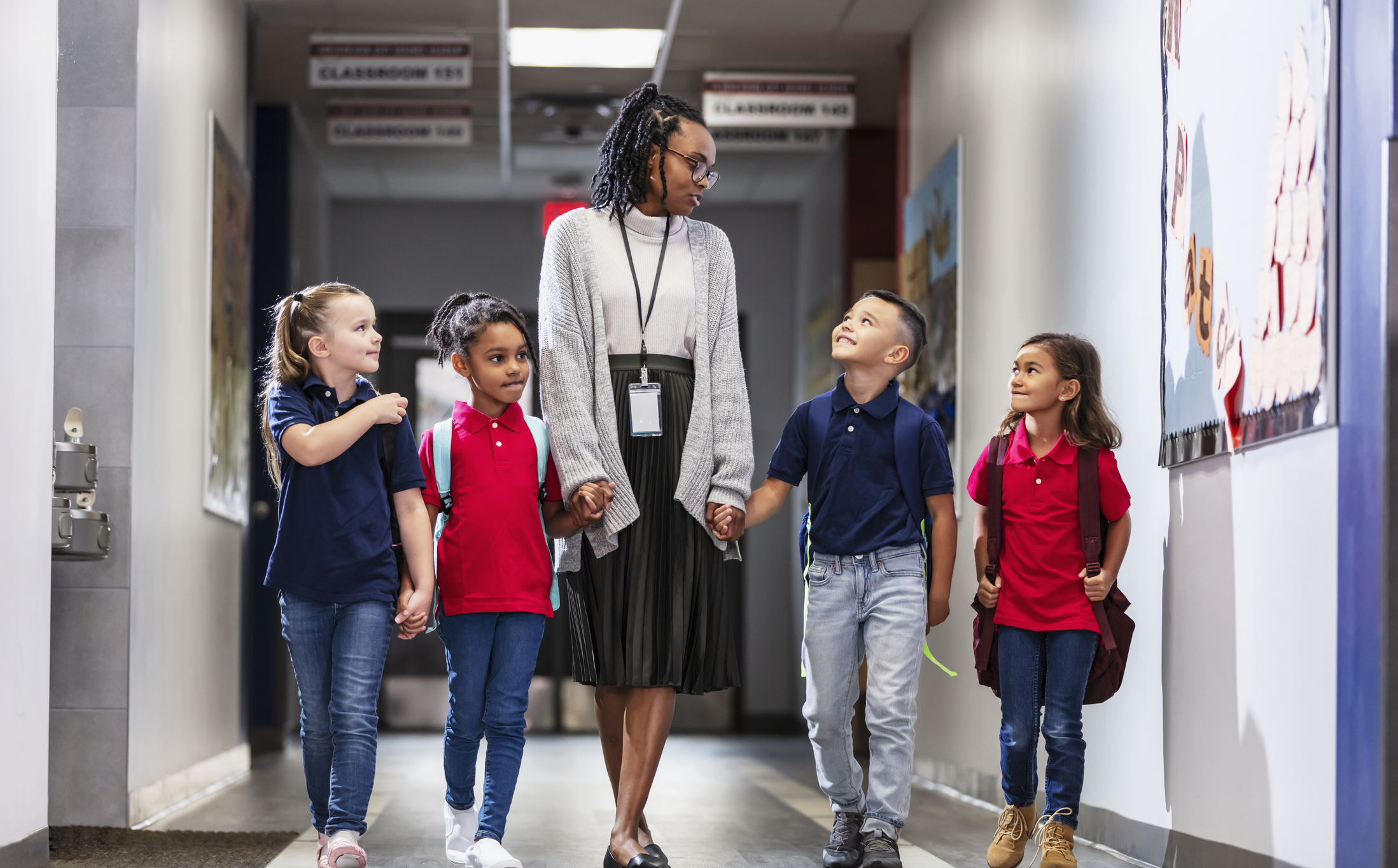 A black elementary school teacher with four multiracial students in a hallway, walking side by side, holding hands. The teacher, a mid adult woman in her 30s, is looking toward an Hispanic boy. The other students are girls. The children are 6 and 7 years old.