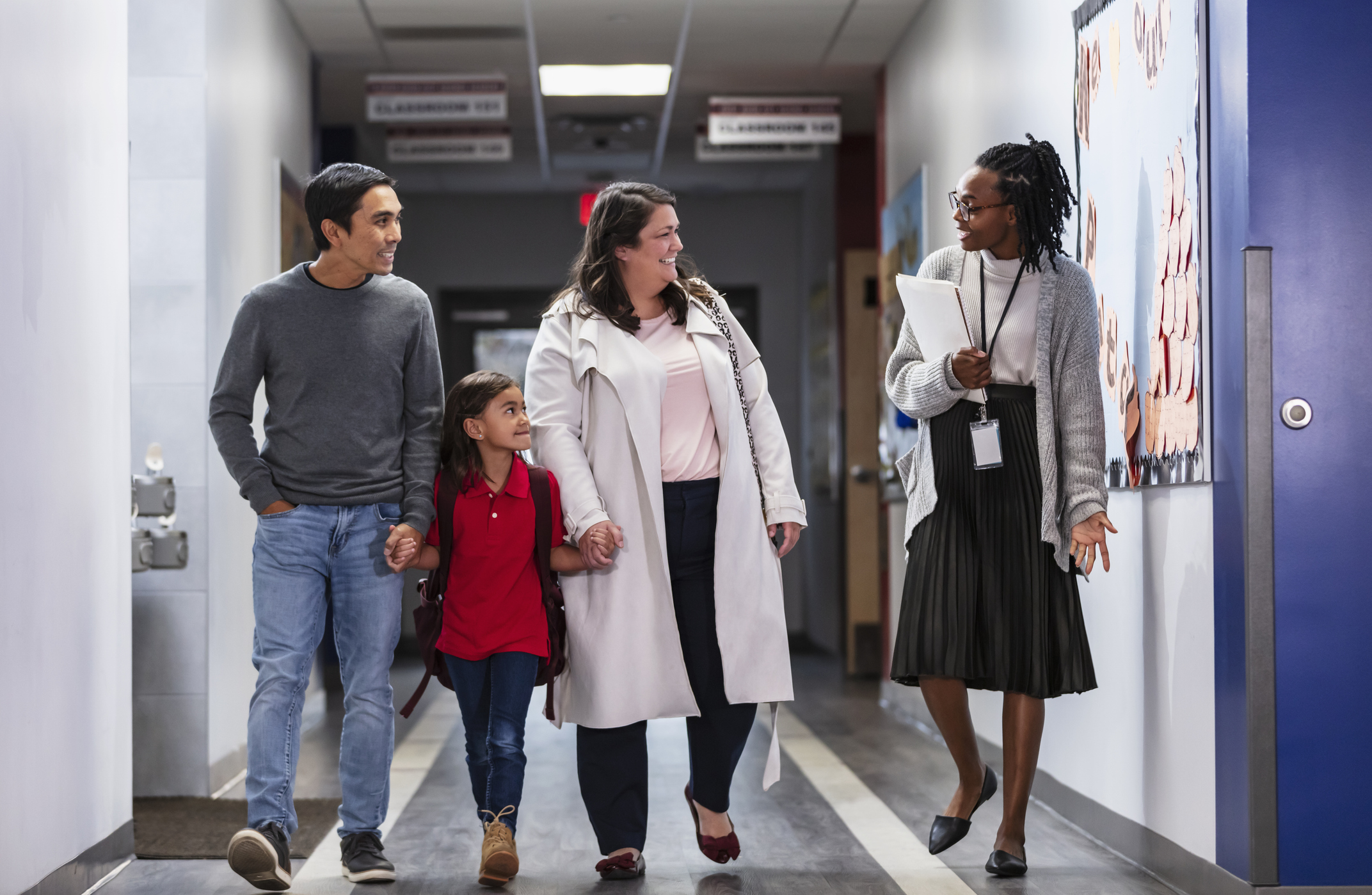 Two parents bringing their daughter to school, talking with an elementary school teacher or principal in the hallway. Perhaps it's the first day of school, or they are going to have a parent-teacher conference. The multiracial girl is 6 years old. Her father is Asian and her mother is Hispanic.