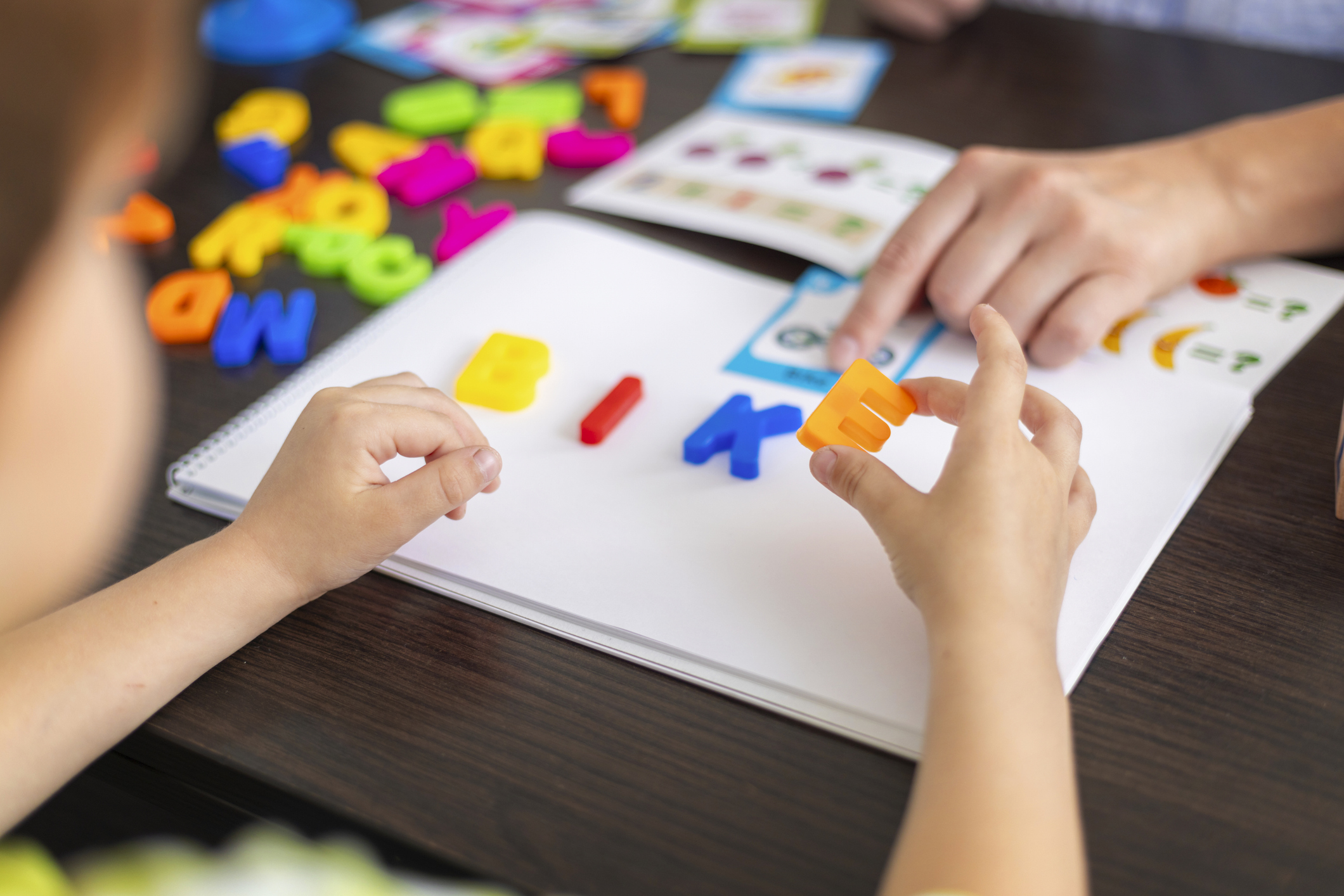 A child learns spelling using colorful plastic letters on a notebook, forming a word while an adult points at an educational activity sheet. Indoor home or classroom learning environment