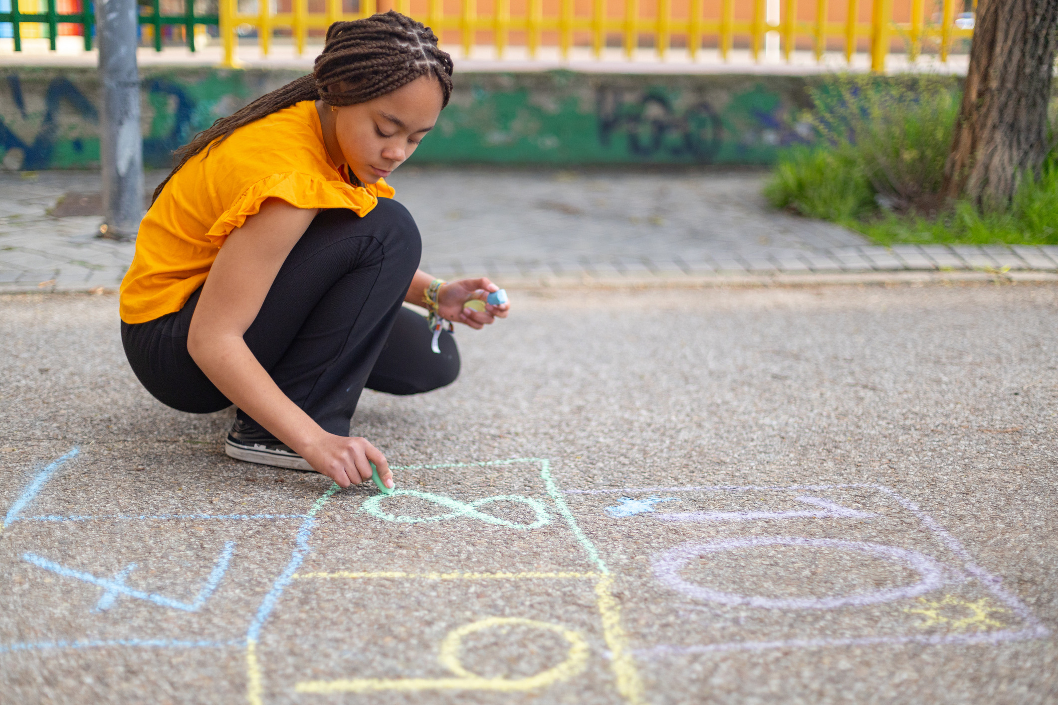 Young girl with braids is crouching down, drawing a hopscotch grid with colorful chalk on the pavement