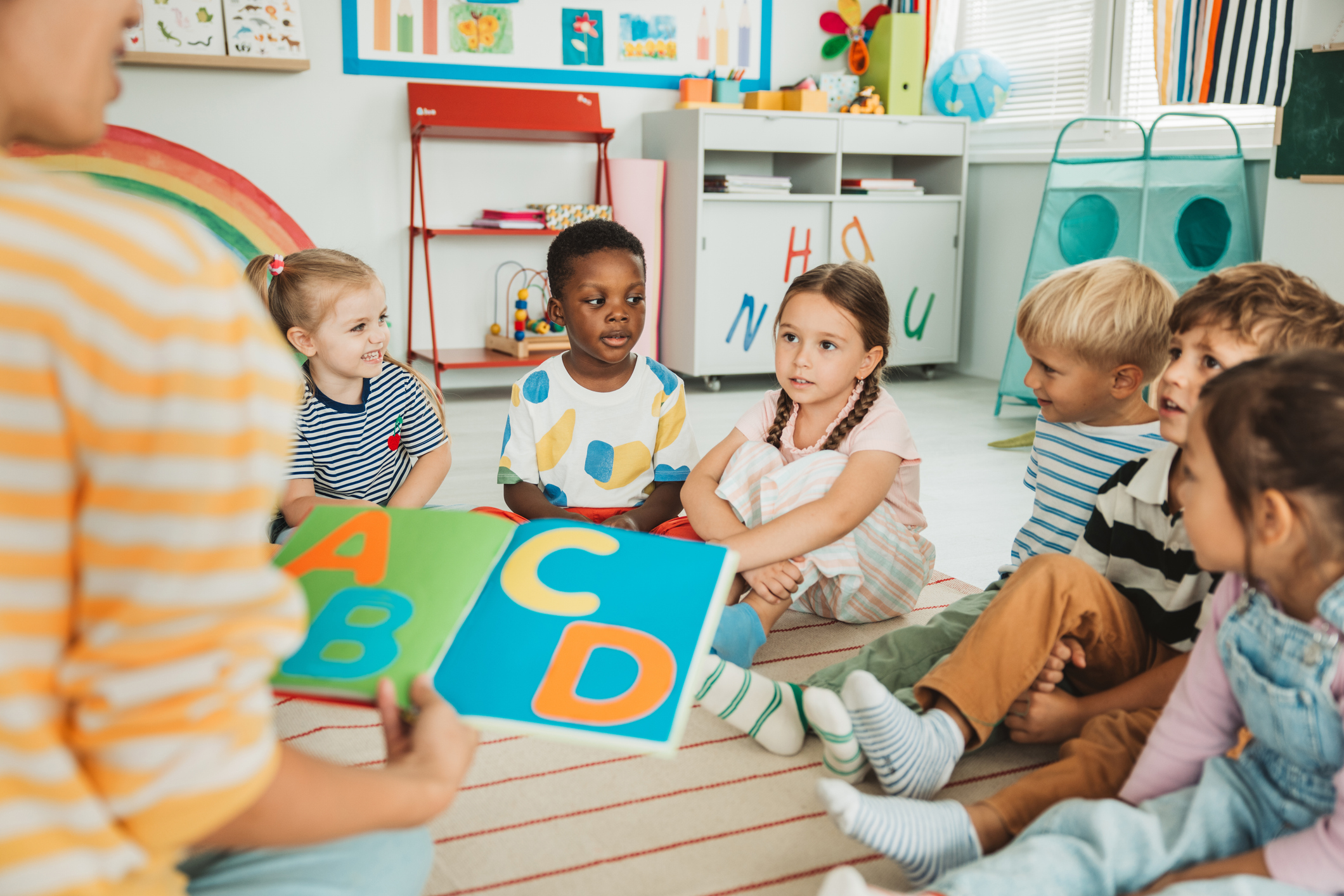 A preschool teacher sits on the floor of her classroom with a small group of students as she reads them a book.