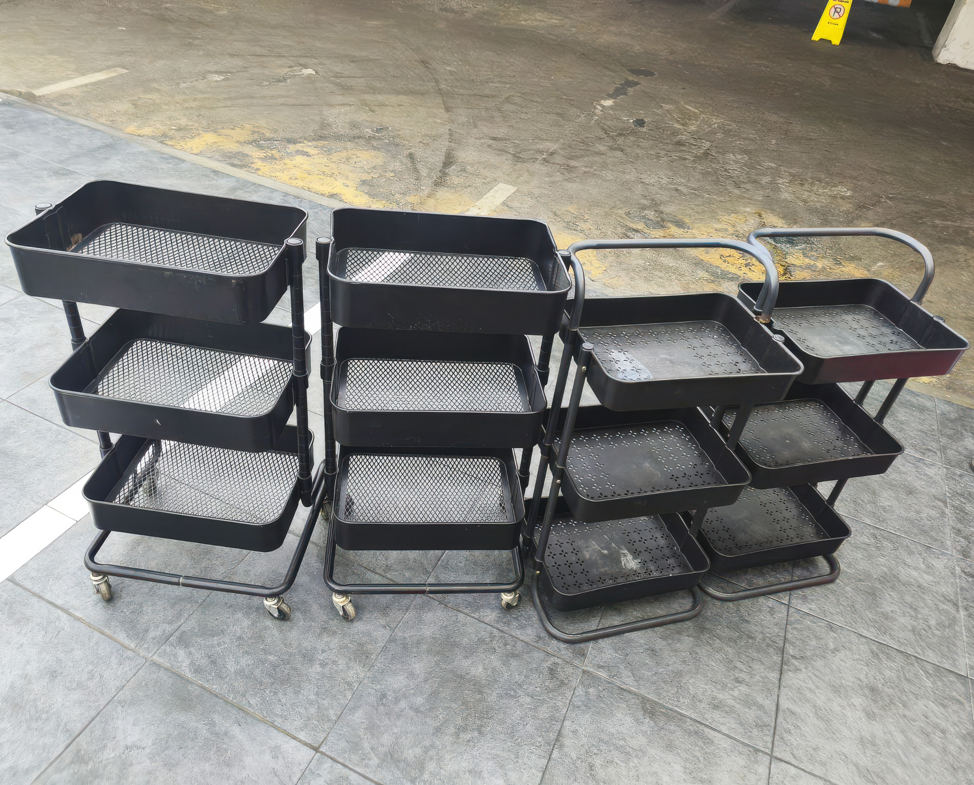 Several black metal rolling utility carts with multi tier trays arranged on tiled floor, used for storage, organization, workshop, or cleaning tasks.