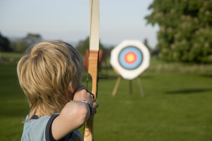 A boy is shown from behind aiming a bow and arrow at a target.