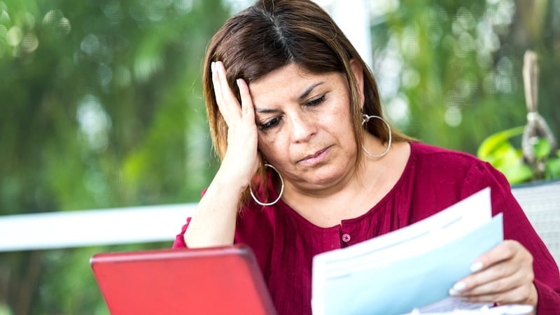 A mature hispanic woman is sitting at the dining table using her tablet to sort her household bills online.She is looking glum as she struggles to sort the family finances. She is holding an invoice .The tablet is surrounded by various bills and debt letters