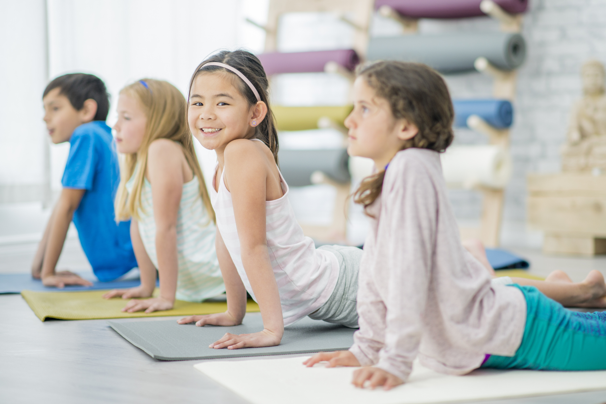 A multi-ethnic group of children are indoors in a health club. They are wearing casual athletic clothing. They are doing a yoga pose while looking serious. One Asian girl is smiling at the camera.