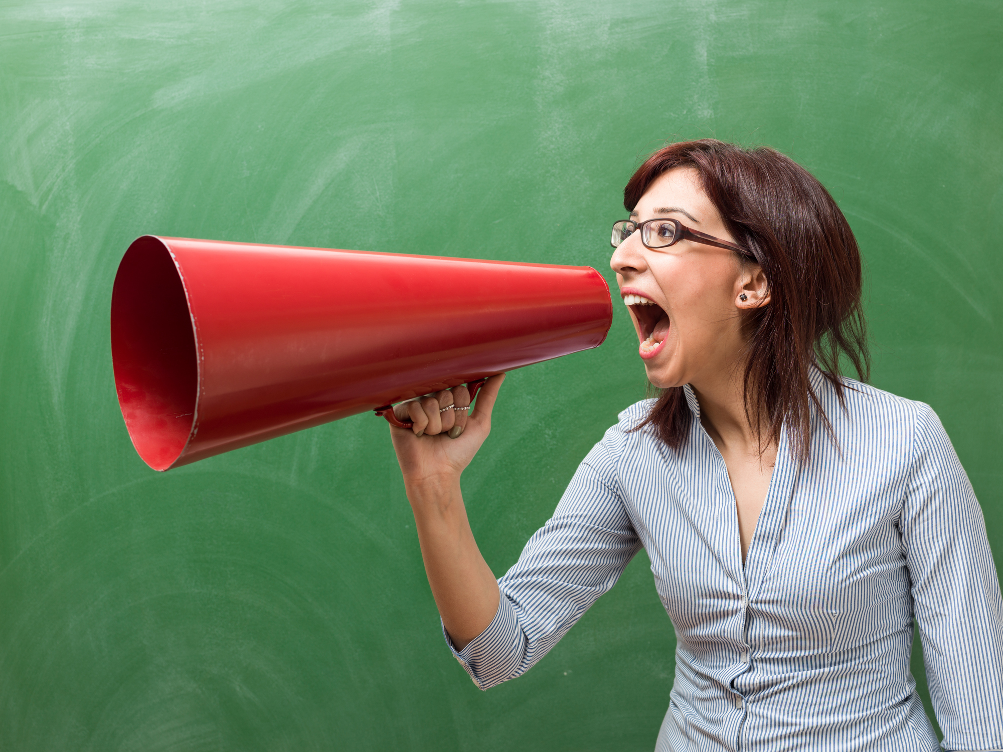 Adult woman wearing a striped shirt shouting through megaphone in front of green chalkboard. She has brown hair and standing on the right side of horizontal frame. The background is dirty blank green blackboard. Shot in studio with medium format camera.