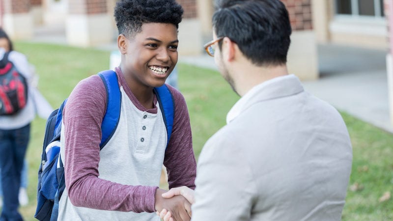 A teenage boy shakes hands with a High School teacher between classes outside his school building. He is receiving congratulations for a job well done.
