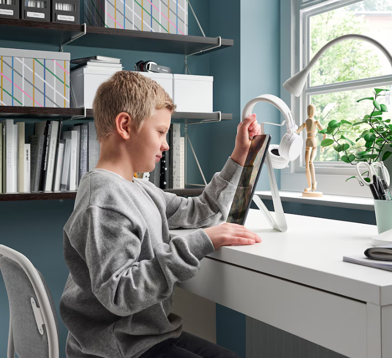 A boy sits at a desk removing a pair of headphones from a white metal stand.