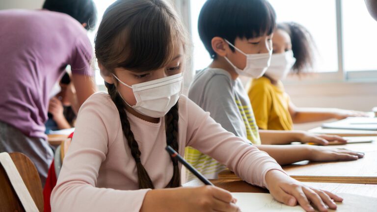 Multiethnic group of school children wearing face coverings sitting at school desks, writing, education, covid 19. Students wearing face masks working in classroom (Multiethnic group of school children wearing face coverings sitting at school desks, w