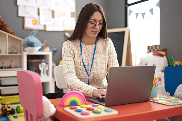 Young beautiful hispanic woman preschool teacher using laptop si
