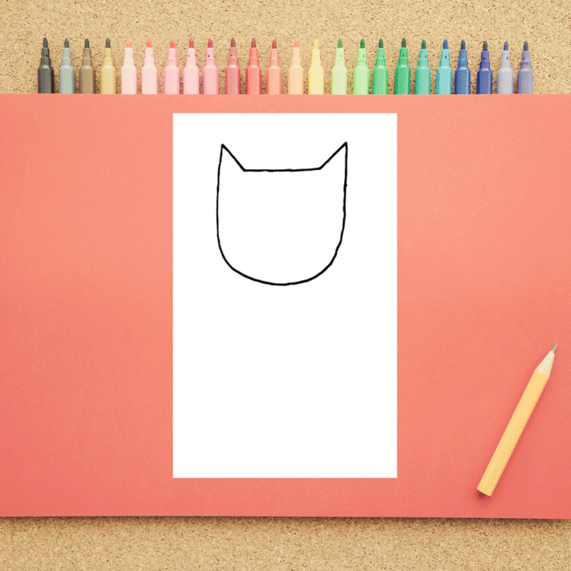 A white piece of paper on a peach background has the beginning sketch of a cat head on it. Markers are shown in the background.
