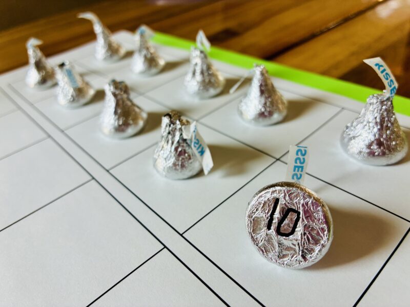10 Hershey chocolate kisses on a tens frame and one kiss has a number on the bottom to help students keep track during a chocolate scavenger hunt
