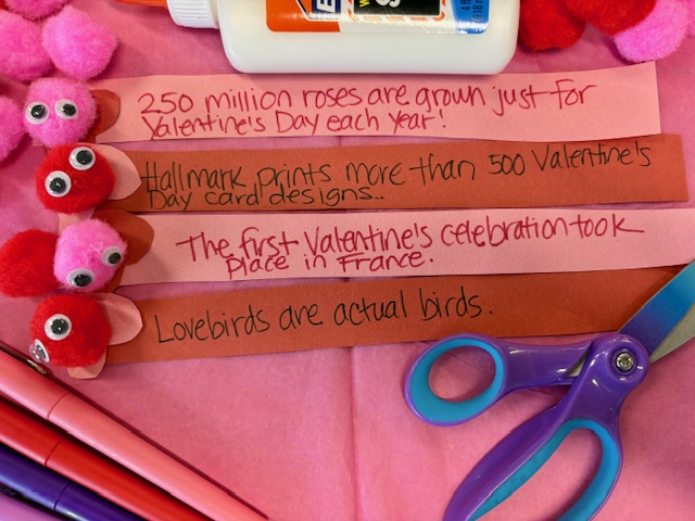 warm-fuzzies Pink and red pom poms with googly eyes and paper feet sit on strips of paper with valentines facts