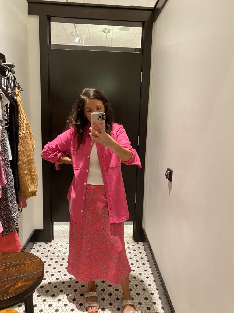 Example of a teacher in a store dressing room wearing a long pink print skirt, pink button down shirt, and sandals as an example of cute teacher outfits.