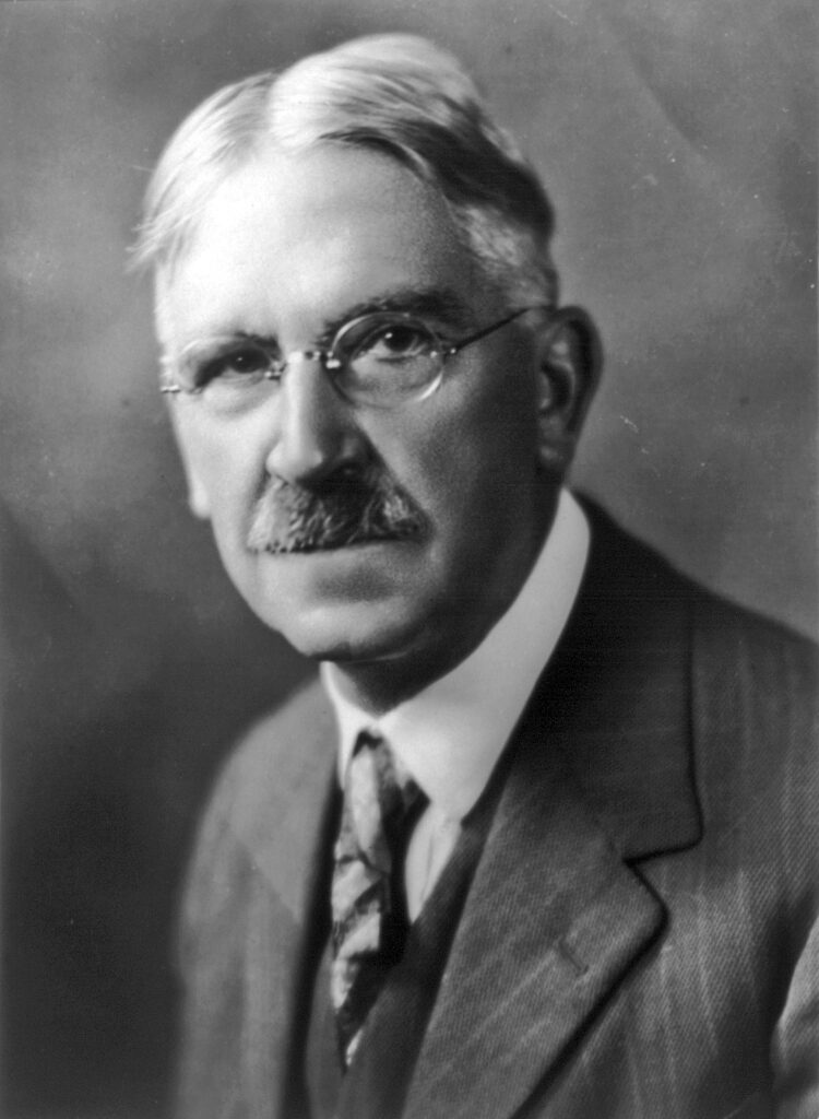 A black and white photo shows an older man in a suit wearing thin wire frame glasses.
