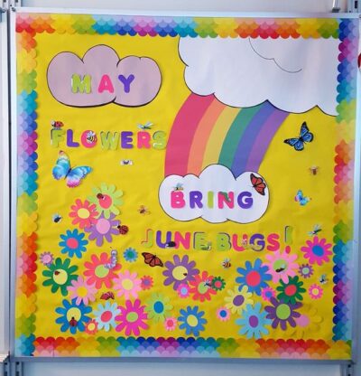 Colorful bulletin board that reads 'May flowers bring June bugs.' Flowers, butterflies, and a rainbow are on the yellow background. 