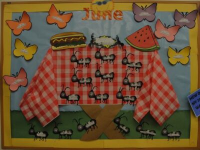 Bulletin board that reads 'June.' A picnic table is in the center with ants, butterflies, and picnic foods on it. 