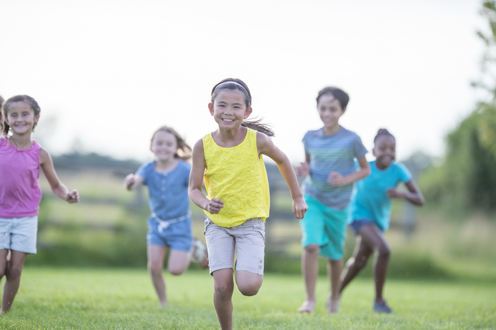 A group of school aged children are seen running across a grassy field on a warm summers day.  They are each dressed casually and are in shorts and t-shirts as they laugh and enjoy the fresh air.