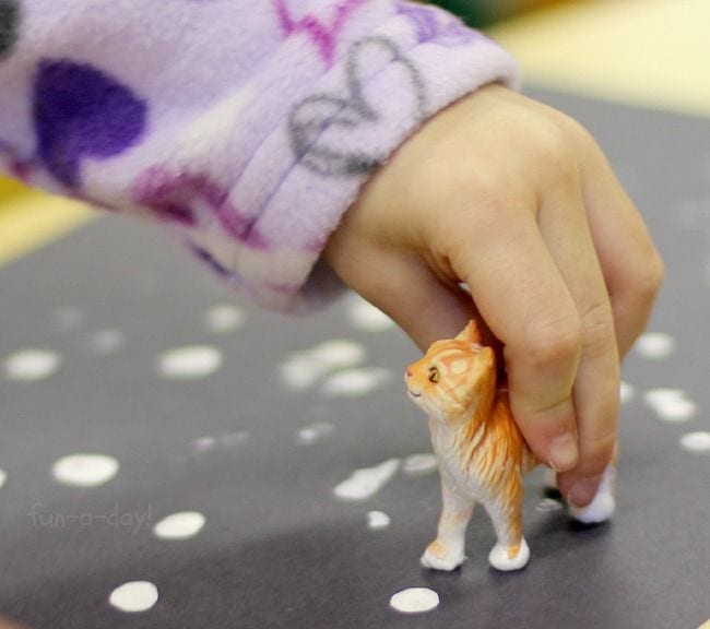 Child's hand holding a plastic cat figuring, using it to make paint white paw prints on a sheet of black paper