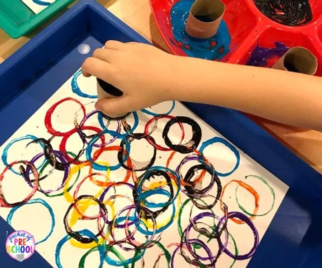 A child's hand is seen making circle prints with a toilet paper roll and paint in this example of kindergarten art projects.