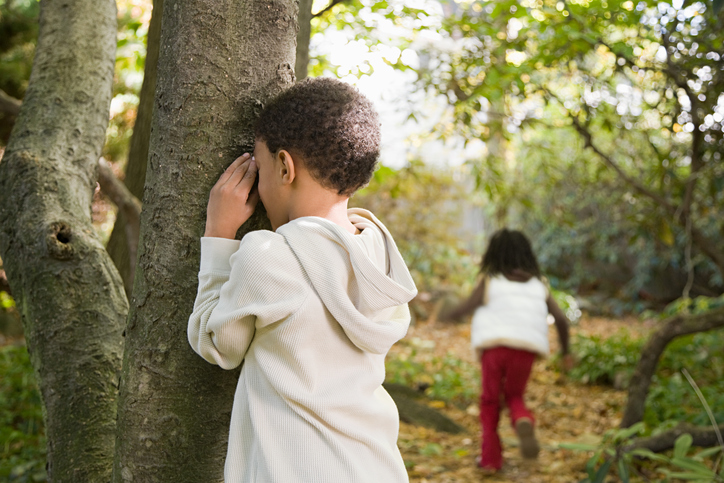 Recess games similar to hide and seek include this one in which a little boy is seen covering his face against a tree while another child runs away in the distance.