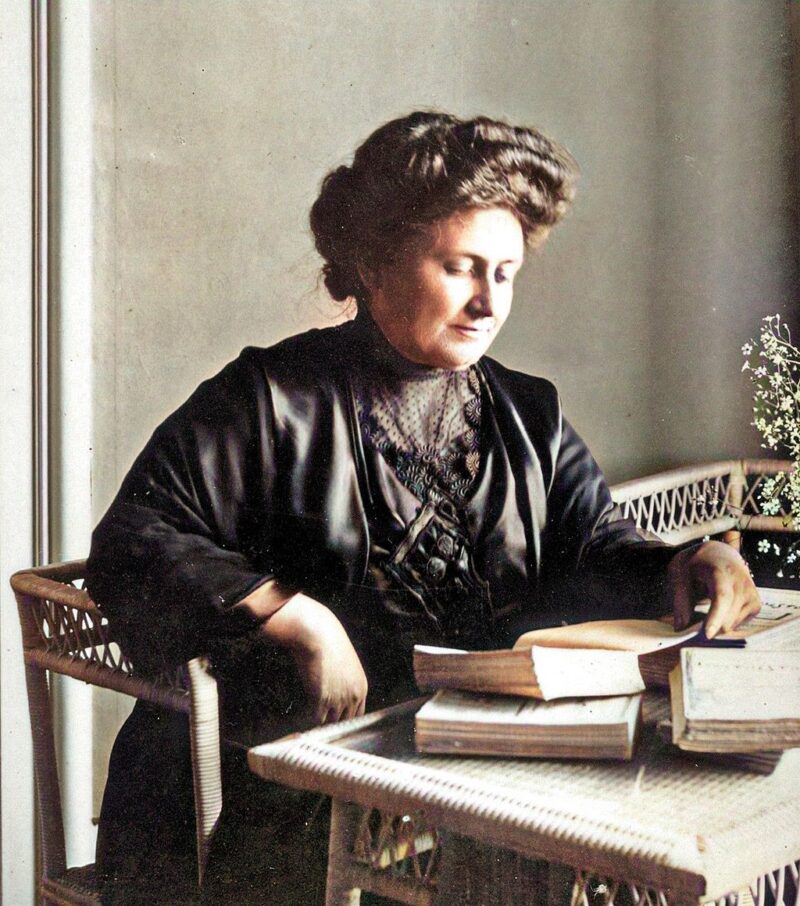 Color portrait of Maria Montessori in 1913 sitting at desk reading book.