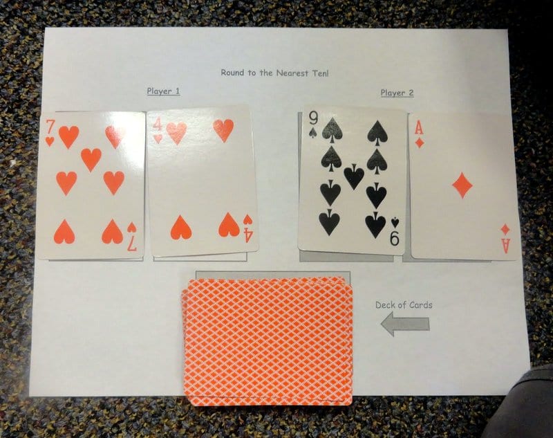 Playing cards laid out on printable worksheet called Round to the Nearest Ten (Math Card Games)
