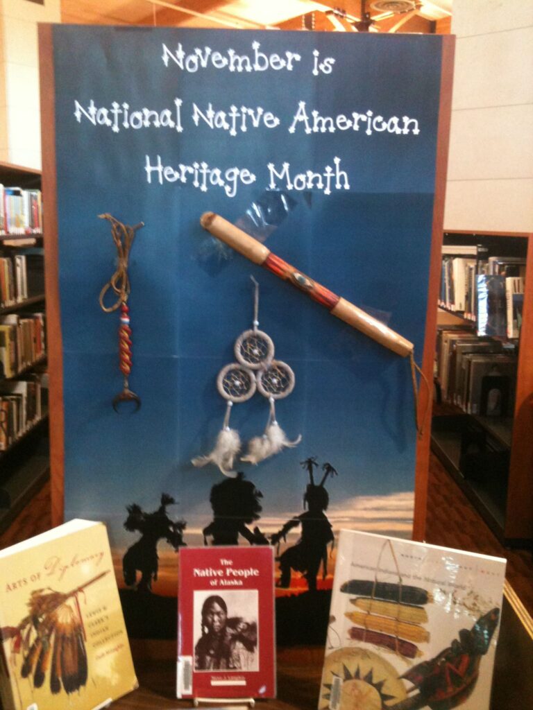 A Board reads "National Native American Heritage Month." A talking stick, dreamcatcher, and other items are attached to the board. Several books on native americans re shown beneath the board. 