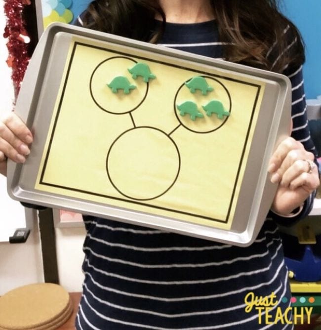 Teacher holding a metal cookie sheet with a number bonds worksheet and dinosaur magnets