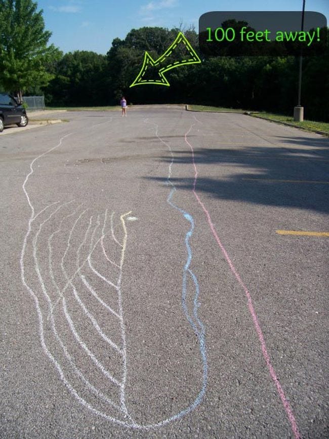 A large whale has been drawn with sidewalk chalk on a black top. There is an arrow and text that reads 100 feet away. 