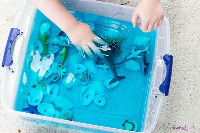 A child's hands are reaching into a bin filled with water, toy sea creatures, shells, etc. (ocean activities)