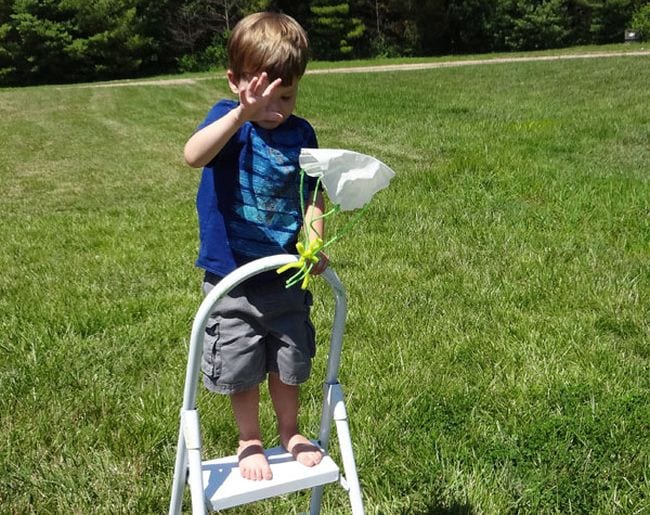 Child standing on a stepladder dropping a toy attached to a paper parachute