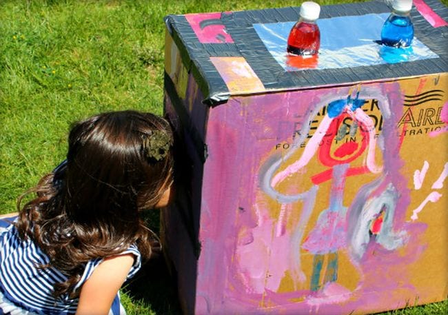 Student peering into a painted cardboard box with red and blue water bottles inserted into the top