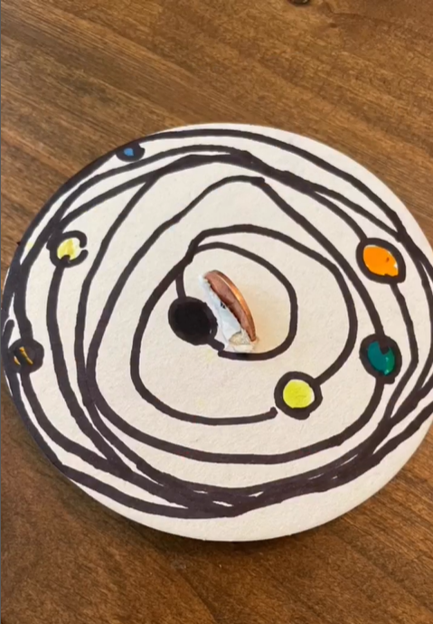 A small white circle has black swirls and circles drawn on it to look like the solar system. A penny is in the middle vertically.