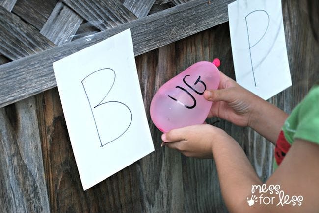 Child holding a pink water balloon labeled with the letters UG next to a paper target labeled B