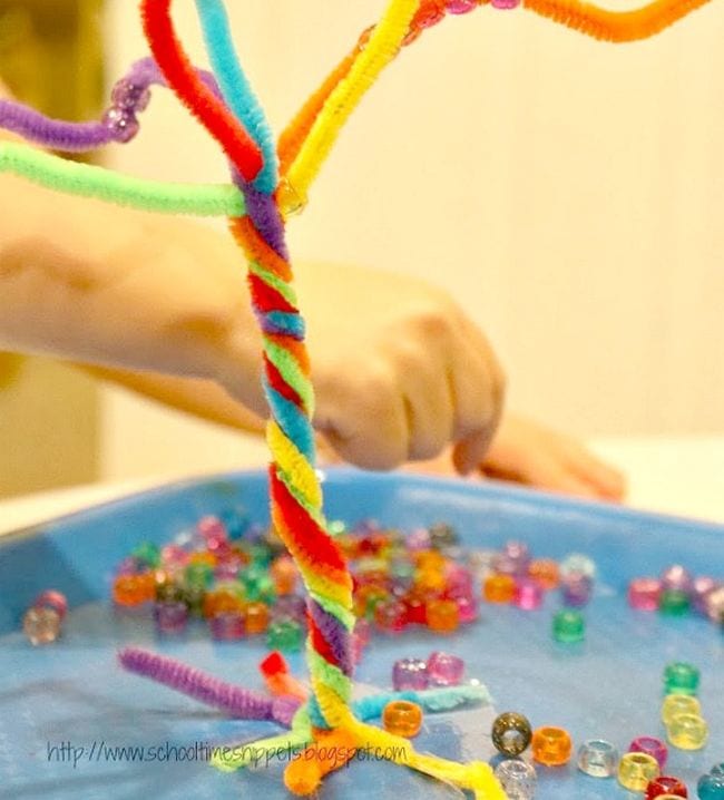 Rainbow pipe cleaners are twisted into a tree shape and a hand is seen stringing beads onto it.