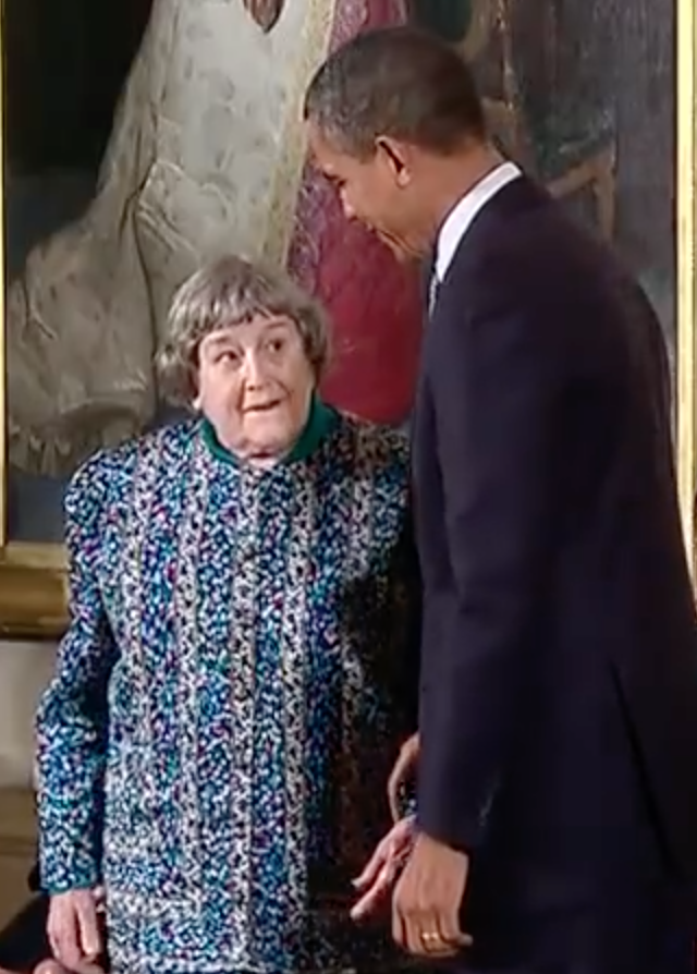 An older woman in a blue dress is seen standing beside President Obama.